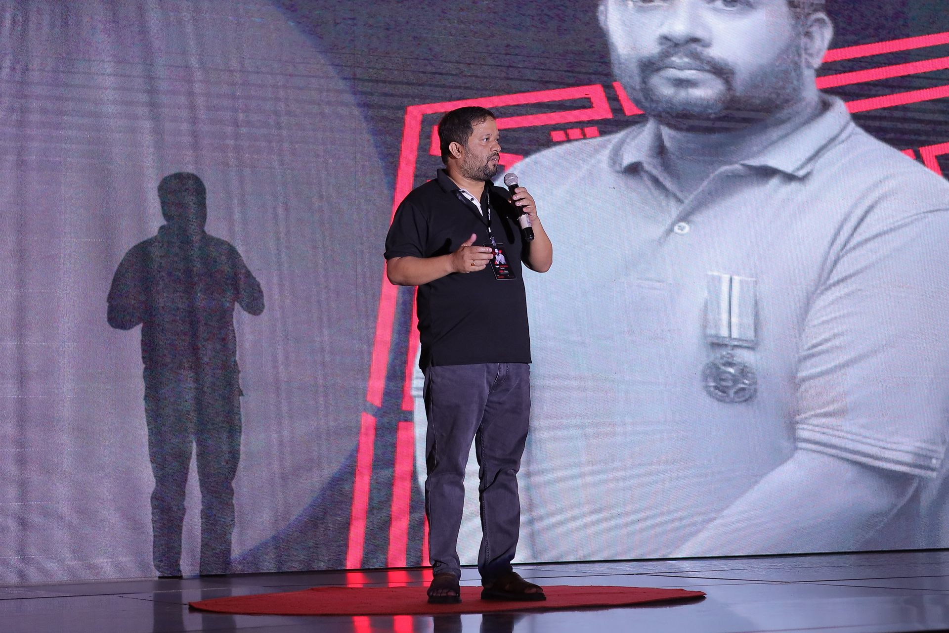 Speaker presenting on the iconic red TEDx dot stage at TEDx Clifton organized by CoinConnect, with large black-and-white speaker portrait projected behind — full TEDx production aesthetic