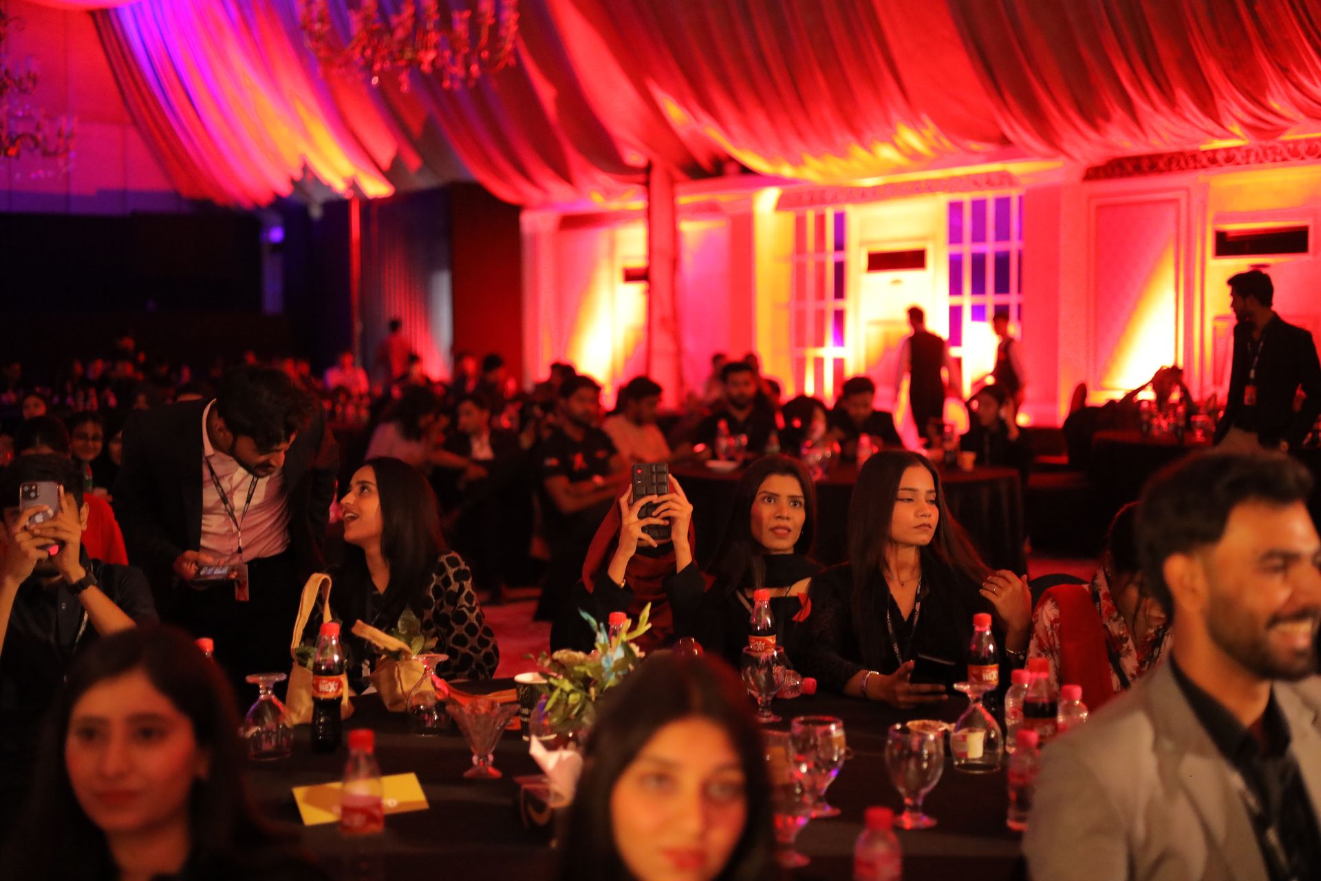 Wide audience shot of the TEDx Clifton venue with red and amber stage lighting, packed banquet seating, and capacity attendance — proving event scale and production quality